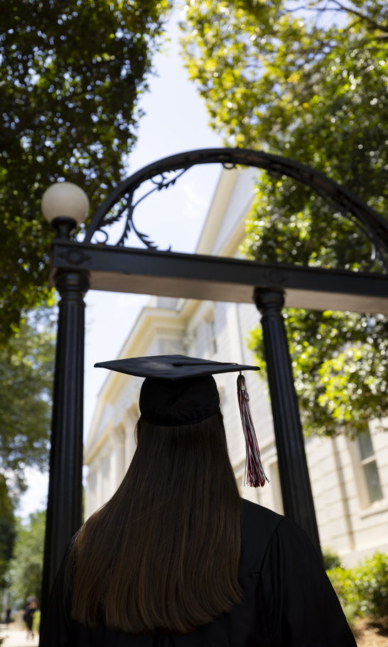 A graduate in cap and gown faces the Arch.