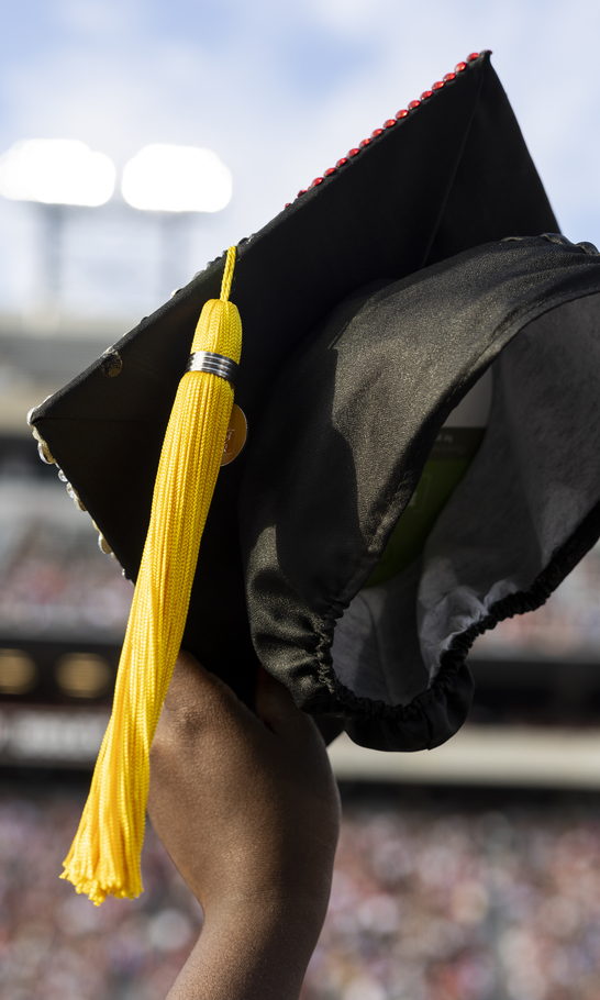 A graduate holds a decorated cap and gold tassel above the crowd at Sanford Stadium.